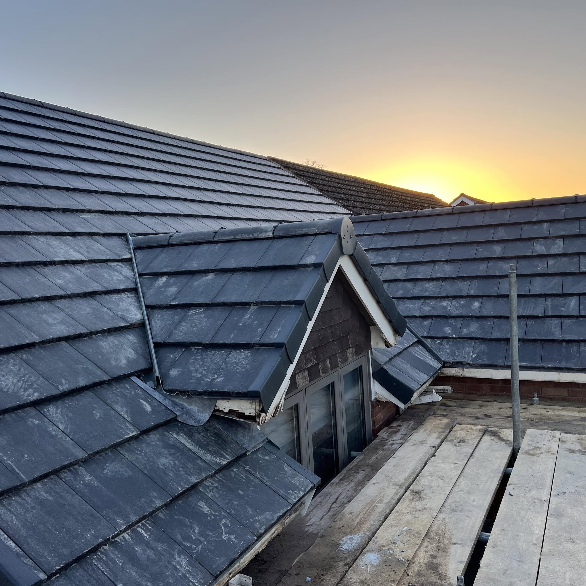A newly tiled slate-grey roof with a dormer at sunset, scaffold boards in the foreground