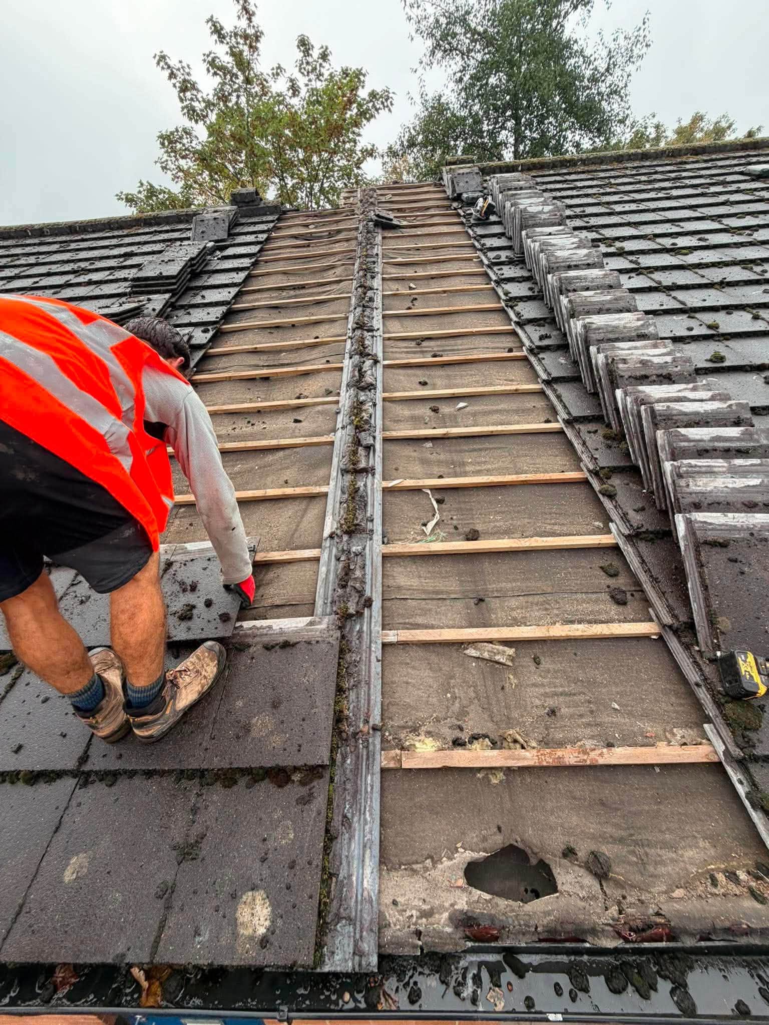 A roofer in a hi-vis vest stripping old tiles back to the battens, ridge tiles stacked beside him