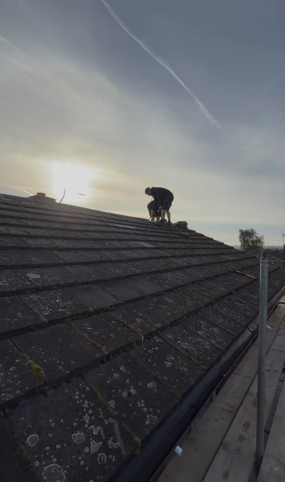 A roofer silhouetted against the sky, working on a tiled roof beside the scaffold rail