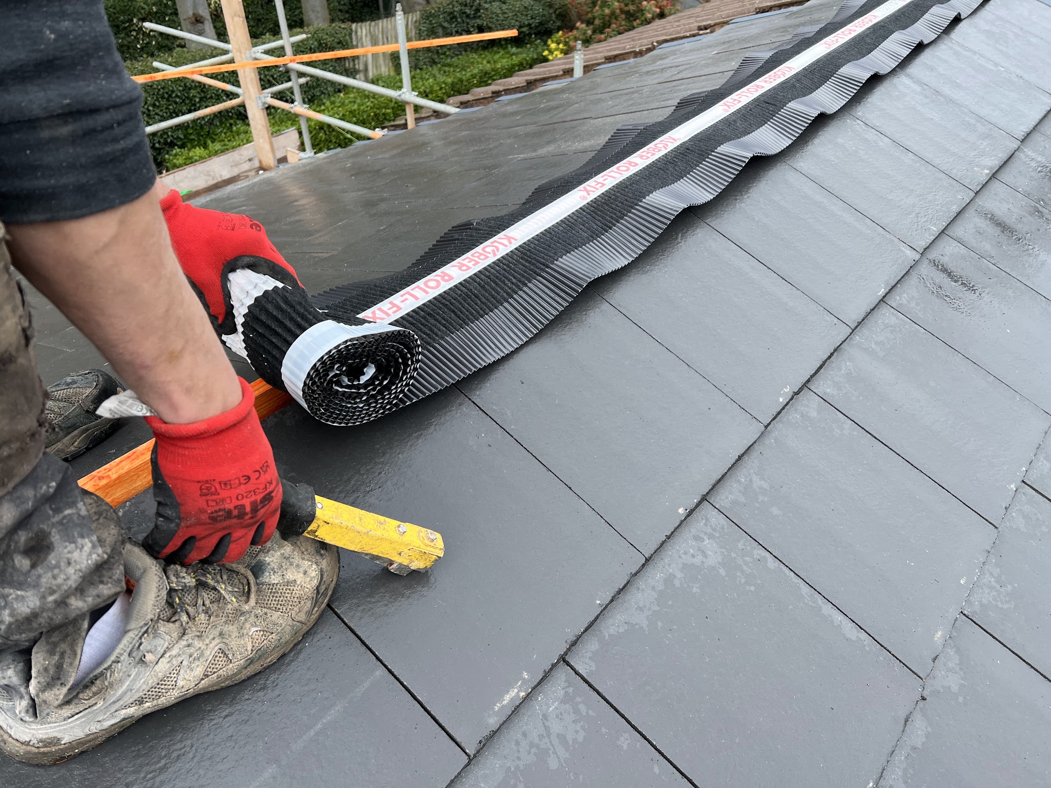 Gloved hands rolling out a dry ridge roll along new slate-grey roof tiles