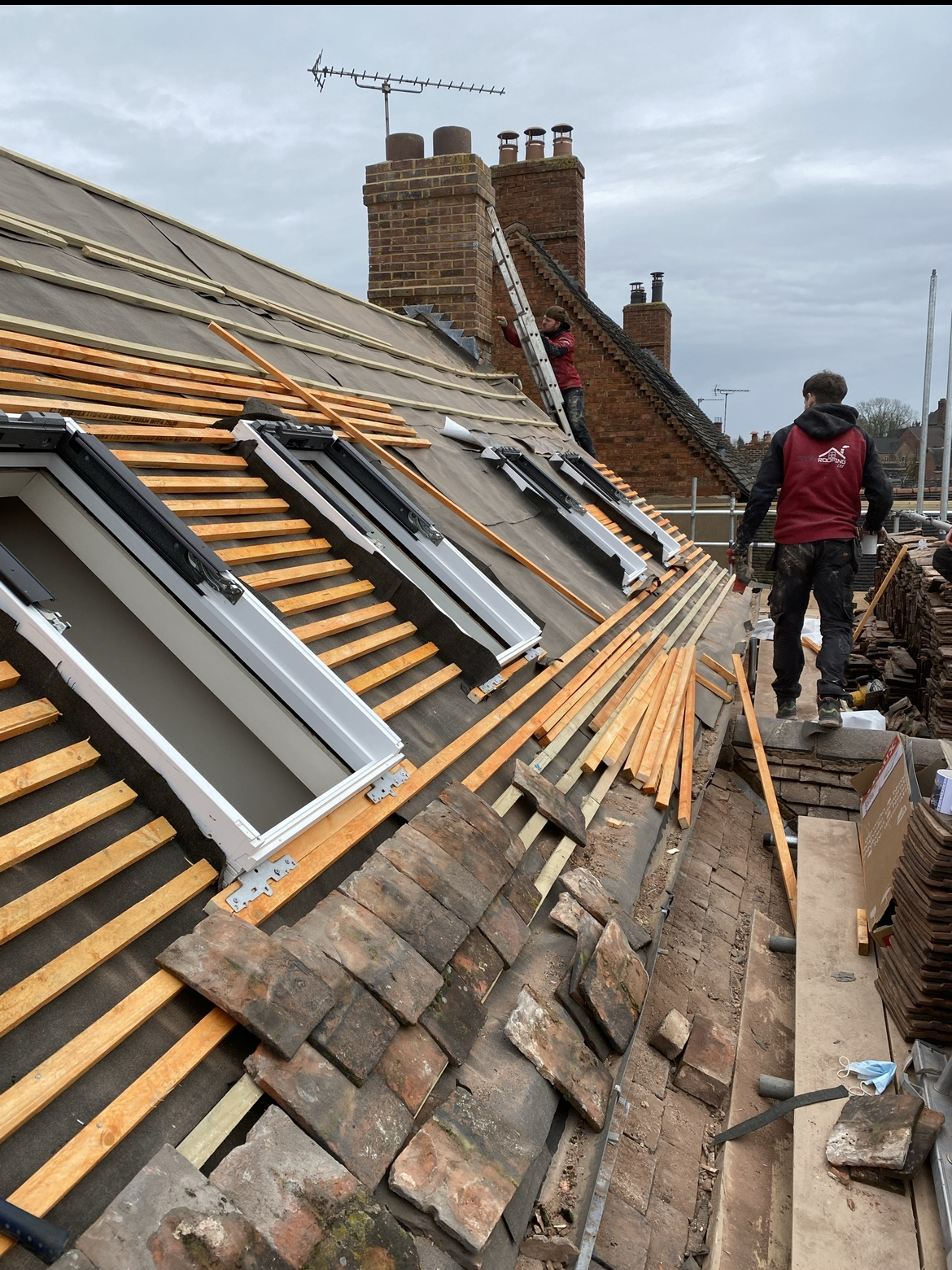 A pitched roof stripped to fresh timber battens around two roof windows, old tiles stacked on the scaffold