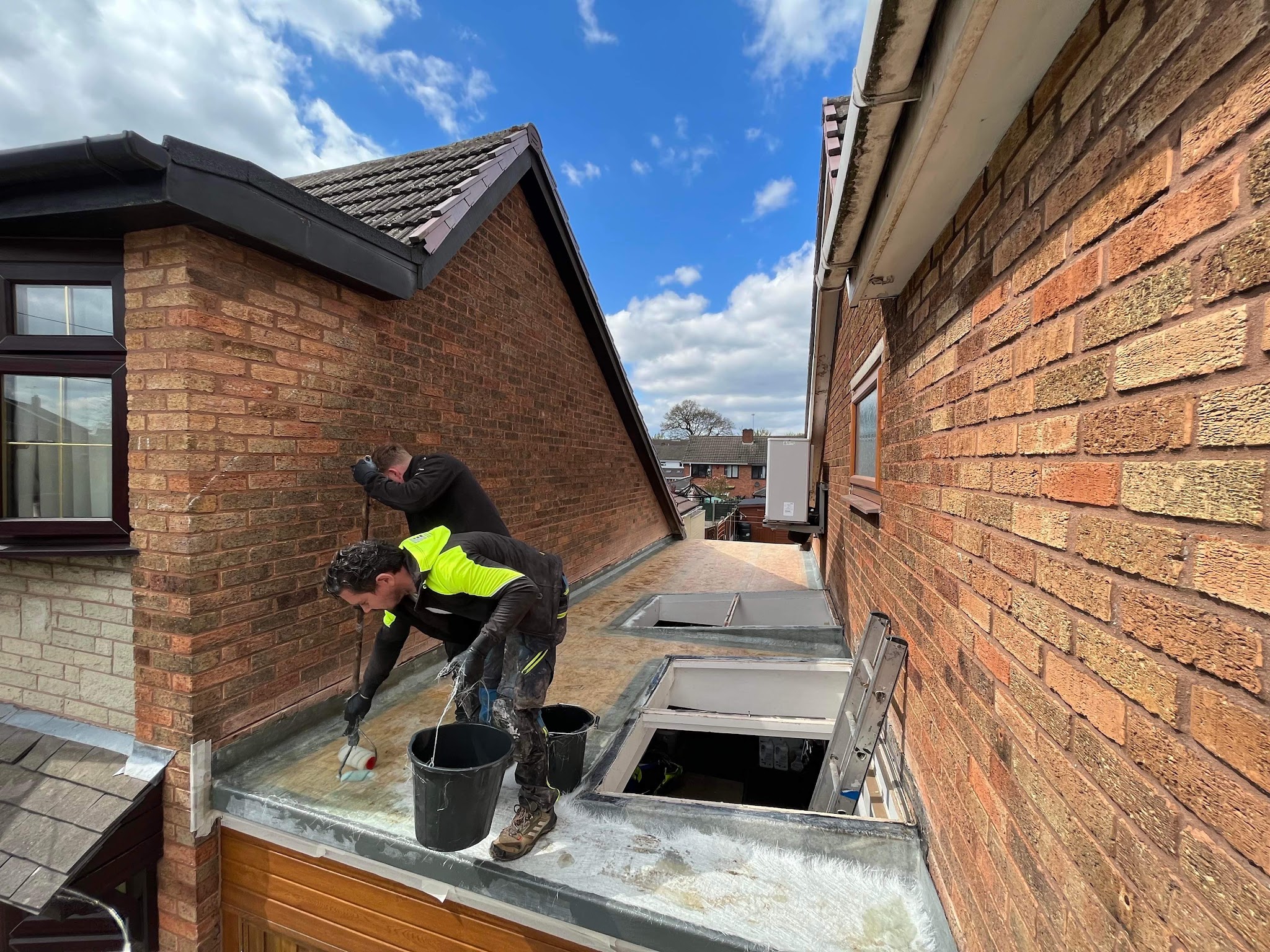 Two roofers working on a flat roof between two houses, with roof lights open