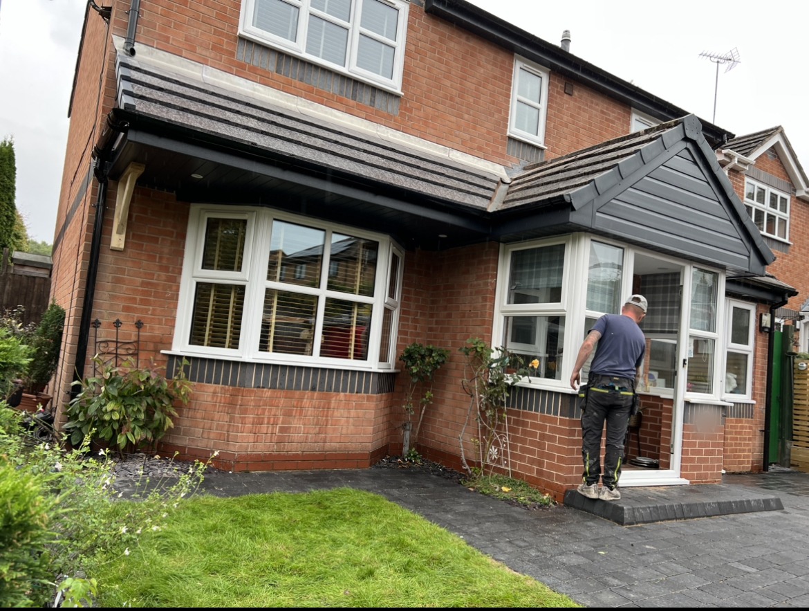 A finished pitched tiled roof over a bay window and porch on a red-brick house, a roofer at the door